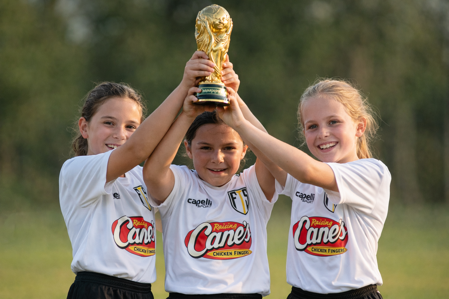 Three young First Touch Soccer campers in FTS jerseys hoisting a World Cup trophy